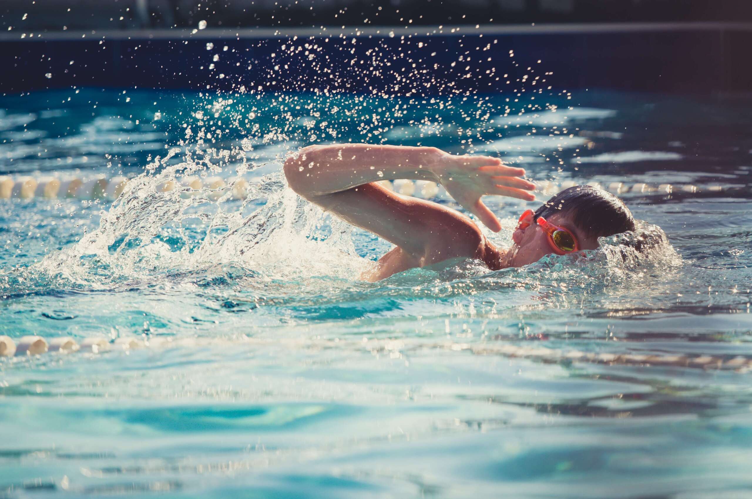 Jugendlicher Schwimmer mit roten Schwimmbrille, der im Wasser beim Brustschwimmen ist, spritzt Wasser auf.