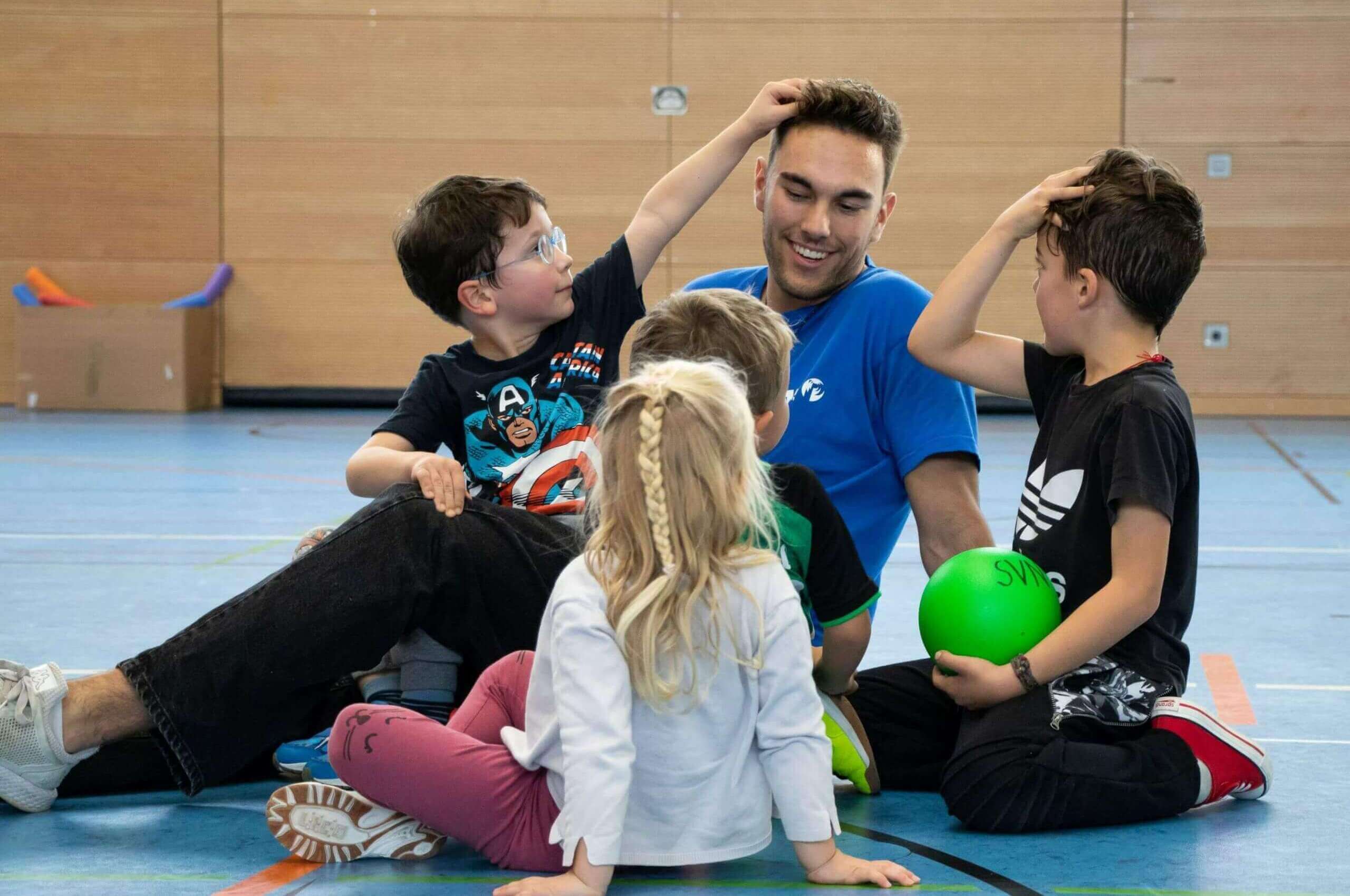 Ein junger Mann sitzt mit vier Kindern auf dem Boden einer Sporthalle, während sie miteinander spielen und lachen.