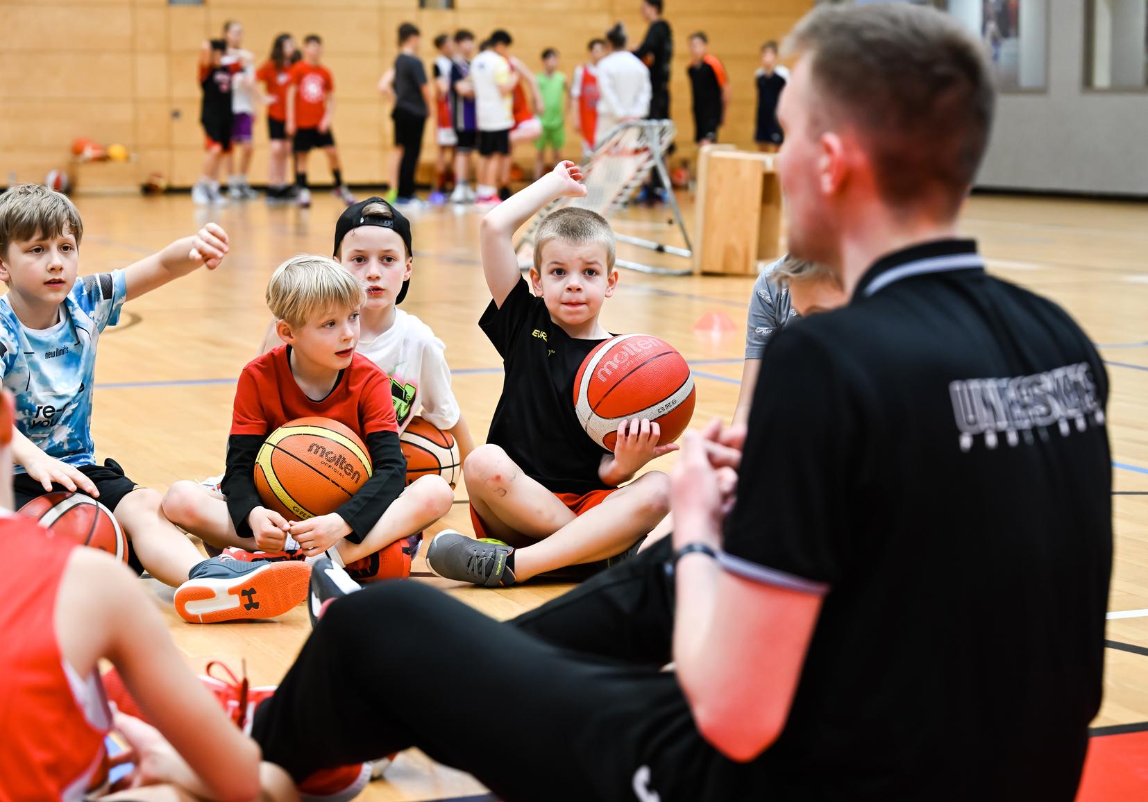 Eine Gruppe von Kindern sitzt auf dem Boden der Sporthalle und hält Basketballs, während ein Trainer ihnen zuschaut.