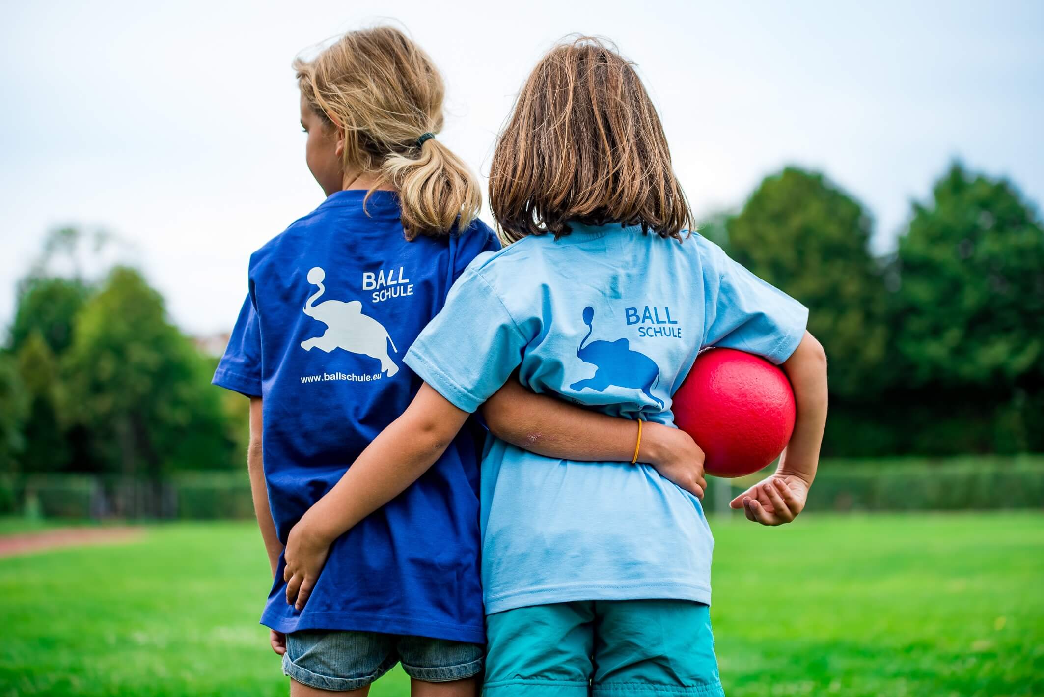 Zwei Mädchen stehen auf einem Sportplatz Arm in Arm. Das rechte Mädchen hält einen roten Ball unter ihrem Arm