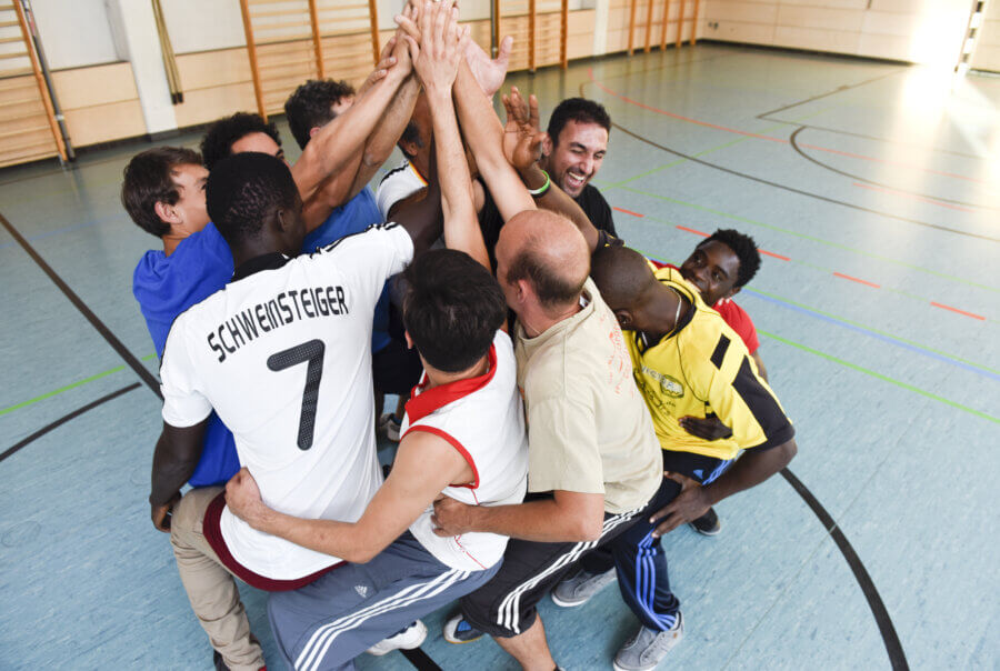 Eine Gruppe Männer sitzen sich gegenseitig auf dem Schoß und bilden einen Kreis. Alle strecken die Hände zur Mitte